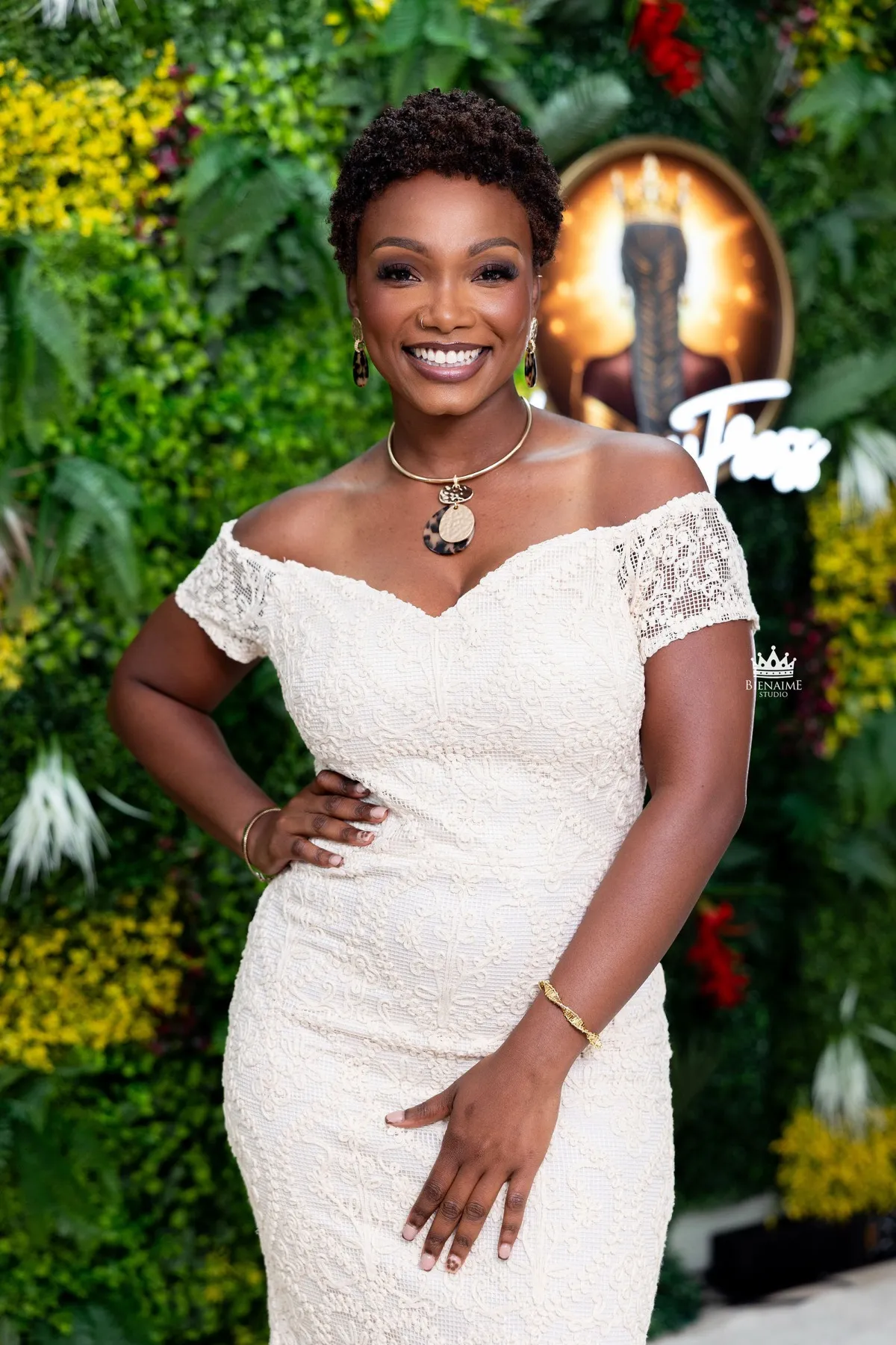 Christelle Bolden, founder of LegacyTress, smiling in an ivory lace dress in front of the salon flower wall